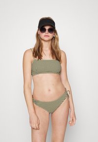 Biquíni verde e branco xadrez com parte de cima sem alças e parte de baixo com laço lateral. Modelo também usa um boné preto e óculos de sol redondos.