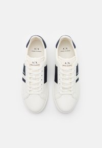 Sneaker bianche con accenti blu navy, materiale sintetico liscio, lacci piatti e un logo visibile sulla linguetta e sui lati.