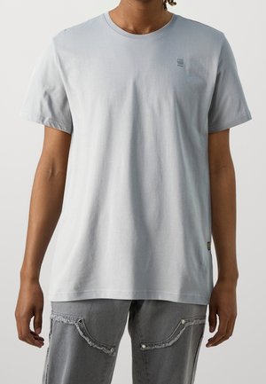 Mężczyzna w prostym jasnoszarym T-shircie z krótkim rękawem i szarych dżinsach z postrzępionymi krawędziami kieszeni, stojący na jednolitym tle.