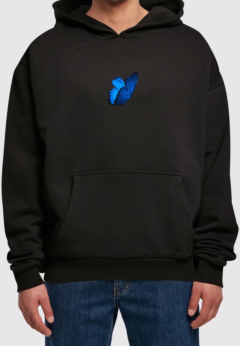 Personne portant un sweat à capuche noir avec une poche avant et un motif de papillon bleu au centre, associé à un jean bleu.