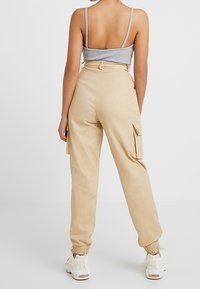 Vrouw in beige hoge taille cargobroek met elastische boorden, een grijs mouwloos shirt en witte sneakers, staande met de handen langs de zij.