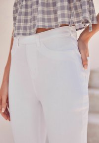 Los jeans de mezclilla blancos presentan un diseño ajustado con una textura suave. Los bolsillos laterales son visibles, complementados por sutiles detalles de costura.