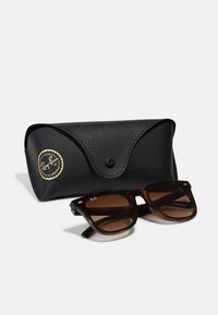 Ochelari de soare cu tortoiseshell maro și lentile maronii, având un cadru dreptunghiular. Însoțiți de o carcasă texturată neagră cu detalii aurii.