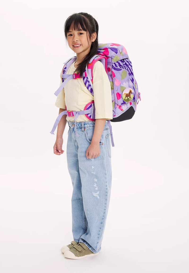 Zaino floreale viola e rosa con spalline regolabili e una toppa rotonda a forma di cavallo, indossato da un bambino con una maglietta gialla chiara e jeans larghi.