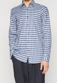 Uomo che indossa una camicia a maniche lunghe a quadri blu e bianchi e pantaloni scuri, con una mano in tasca.