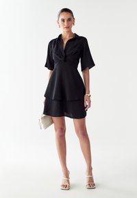 Vestido negro con cintura ceñida, mangas cortas y falda en capas. Textura suave, diseño minimalista, combinado con un pequeño bolso de color claro.