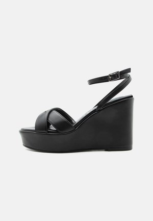 Black wedge sandal with platform sole, two wide crisscross straps over toes, and thin ankle strap with silver buckle.