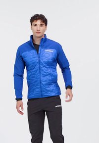 Blauwe geïsoleerde jas met een gequiltde textuur, hoge kraag, volledige rits en zwarte accenten; gedragen met zwarte sportbroeken.