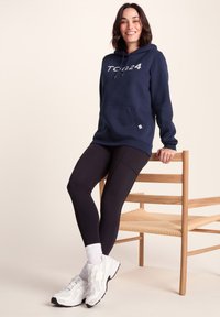 Navy hoodie with "TCB24" in silver, front pocket, paired with black leggings and white athletic shoes, seated on a wooden chair.