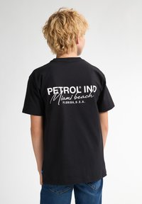 Černé bavlněné tričko s bílým textem na zádech: "PETROL IND Miami beach FLORIDA, U.S.A." Jednoduchý design, kulatý výstřih, krátké rukávy.