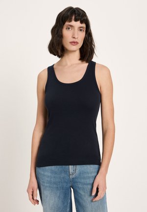 Frau mit dunklem, welligem Haar, die ein ärmelloses schwarzes Tanktop und blaue Jeans trägt, vor einem schlichten hellen Hintergrund stehend.