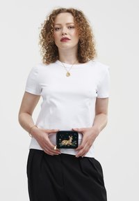 Svart påse med en broderad design av en hjort på en mörk bakgrund, hålls av en person som bär en vit skjorta och svarta byxor.