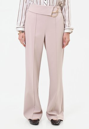 Pantalon beige taille haute avec une large ceinture, plis devant, détail chaîne dorée, porté avec une chemise rayée et des chaussures noires à bout carré.