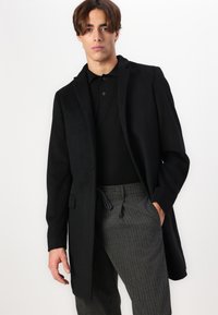 Cappotto lungo nero realizzato in tessuto testurizzato, con colletto a rever, chiusura con un bottone e tasche laterali, abbinato a pantaloni grigi a righe.