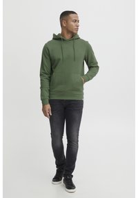Green hoodie with a front pocket, adjustable drawstring hood, and ribbed cuffs, paired with slim-fit dark jeans and black shoes.
