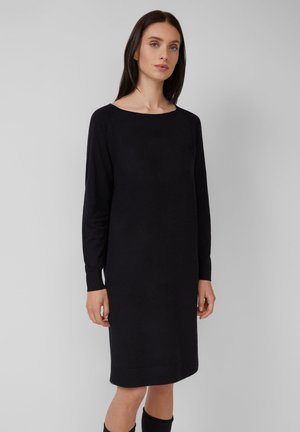 Femme aux longs cheveux noirs portant une robe noire simple à manches longues, arrivant aux genoux, se tenant devant un fond gris clair uni.
