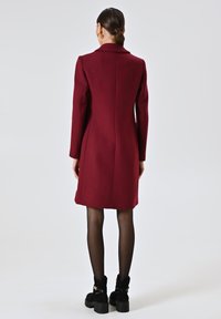 Bordeaux wollen coat met een op maat gemaakt ontwerp, met een geknipt kraag, lange mouwen en een middellange zoom. G gedragen met zwarte enkellaarsjes en panty’s.