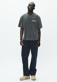 Szary, oversize bawełniany T-shirt z logo "Double V", zestawiony z ciemnoniebieskimi spodniami o szerokich nogawkach oraz beżowym obuwiem. Gładkie tło.
