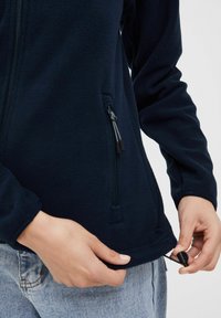Giacca in pile blu navale con tasca con zip, orlo con cordino elastico e maniche aderenti, mostrata da vicino rispetto alle mani, mettendo in evidenza la texture e i dettagli.