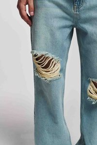 Käsi puhkeb reie peale, kandes helesiniseid rebendiga teksaseid, millel on suured, kulunud augud, mis näitavad nahaala mõlemal jalal.