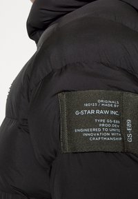 Černá péřová bunda s prošívanou texturou, obsahující látkovou nášivku v olivově zelené barvě přišitou na levé části ramene.