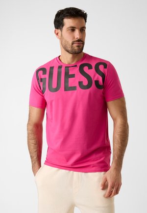 Uomo che indossa una maglietta rosa acceso con grandi lettere nere "GUESS" sul petto, abbinata a pantaloni beige chiaro.
