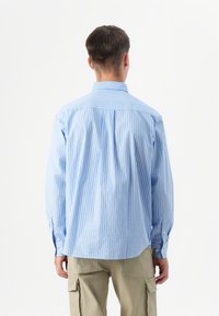 Joven vestido con una camisa a rayas blanca y azul claro y pantalones caqui, visto desde atrás contra un fondo blanco.