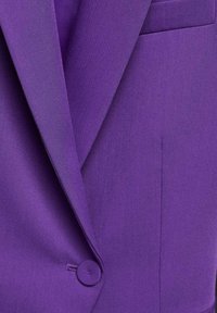 Blazer violet avec un tissu lisse et texturé, des revers nets, une fermeture à un bouton et un détail de poche avant.