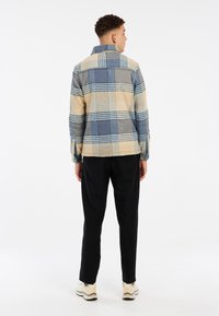 Veste à carreaux en beige, bleu et gris. Tissu doux, poignets à boutons, col et coupe décontractée. Portée avec un pantalon noir et des baskets.