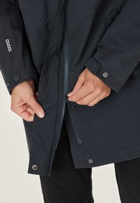 Marineblauwe waterdichte jas met een ritssluiting aan de voorkant en klittenbandsluiting. Voorzien van drukknoopsluitingen op de mouwen en een gladde textuur.