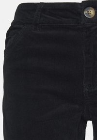 Närbild av svarta kordflabbyxor som visar detalj av midjeband, knapp, bältesöglor och framfickor.