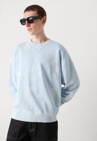 Sudadera azul claro con una textura suave, con un sutil patrón de nubes blancas, cuello redondo y mangas sueltas. Gafas de sol negras.