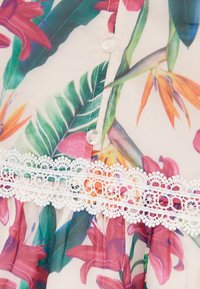 Blouse fleurie avec un imprimé coloré de fleurs et de feuilles sur un tissu léger, agrémentée d'une finition en dentelle blanche le long du bas.