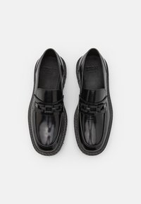 Svart slip-on sko med en glanset finish, med en dekorativ kjedeakcent over toppen og en teksturert såle.