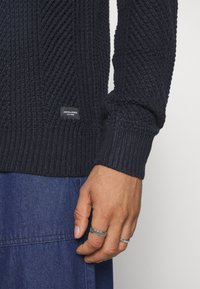Navy ribbed knit sweater with textured design, featuring a small logo tag at the hem. Cuff and body have consistent ribbing detail.