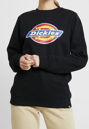 Personne portant un sweat-shirt noir avec un grand logo Dickies coloré sur la poitrine et un pantalon beige, les mains croisées de manière détendue.