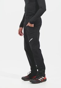 Osoba oblečená v černých outdoorových kalhotách s nápisem "TERREX" na stehně, černém dlouhém rukávu a černo-červených turistických botách značky Adidas.