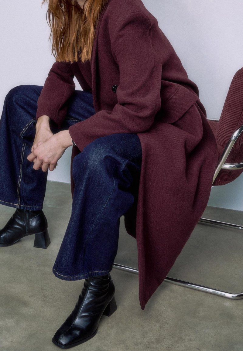 Personne assise sur une chaise en métal portant un long manteau bordeaux, un jean large bleu foncé et des bottines noires à talons, les mains jointes devant.