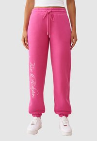 Roze joggingbroek met een koord in de taille, elastische boorden en een glanzend wit tekstontwerp langs het linkerbeen. Gecombineerd met witte sneakers.