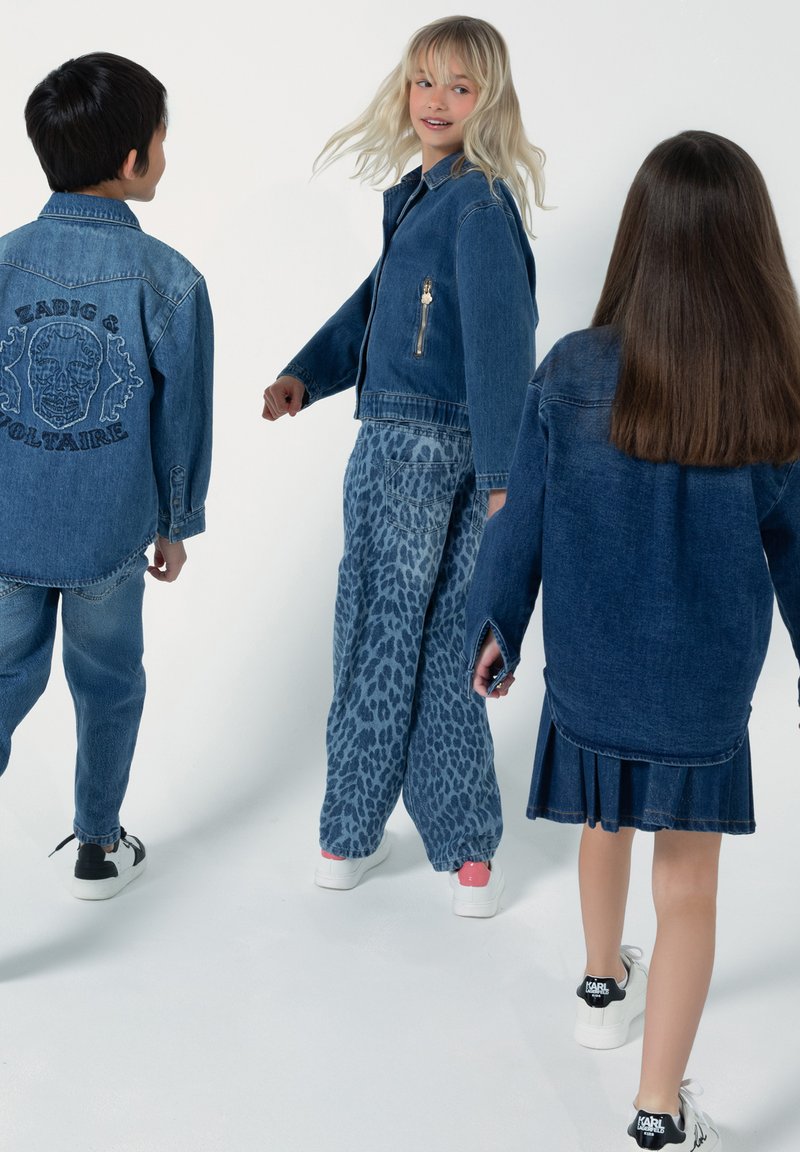 Tre bambini in abiti di denim: una giacca blu chiaro, pantaloni con stampa leopardata e un vestito blu scuro con orlo plissettato. Indossano scarpe da ginnastica.