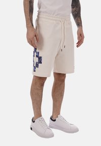 Homme portant un short beige à cordon avec une croix bleue à motifs sur le côté gauche, des baskets blanches et un t-shirt blanc, montrant ses bras tatoués.