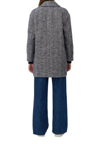 Frau in einem grauen Fischgrät-Muster-Mantel, weit geschnittenen blauen Jeans und weißen Schuhen, von hinten vor einem weißen Hintergrund gezeigt.