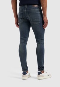 Jean skinny bleu foncé avec une texture délavée, comprenant deux poches arrière et un patch en cuir à la taille, porté avec des baskets blanches.