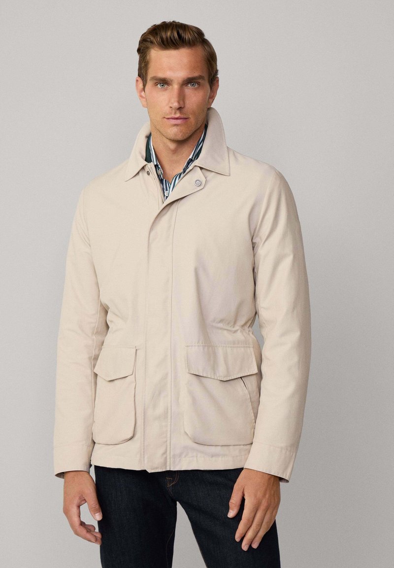 Beige vandtæt jakke med krave, to frontlomme og knaplukning. Bæres over en stribet skjorte, stoffet ser glat ud.
