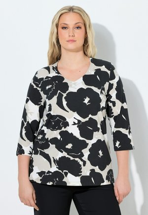 Kvinde med langt blondt hår iført en sort, hvid og grå blomstermønstrede V-hals trøje med 3/4-ærmer mod en hvid baggrund.