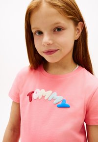 Νεαρή κοπέλα με μακριά κόκκινα μαλλιά που φοράει ροζ μπλουζάκι με 3D γράμματα που σχηματίζουν τη λέξη "Tommy" σε κόκκινες, λευκές, ανοιχτό μπλε και μπλε αποχρώσεις στο στήθος.
