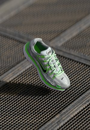 Chaussure de sport en mesh gris avec des accents verts. Présente une semelle texturée, des détails réfléchissants et un logo noir proéminent sur le côté.