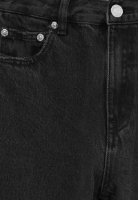 Jean en denim noir en gros plan montrant la poche avant, la boucle de ceinture, les rivets en métal et la fermeture par bouton avec des détails de couture visibles.