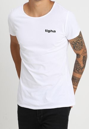 Homme portant un T-shirt blanc ajusté avec le logo "tigha" sur la poitrine gauche et une manche de tatouage noire sur le bras droit, associé à un pantalon noir.