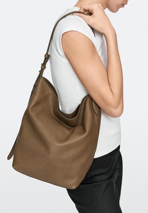 Person in weißem Hemd hält eine große braune Lederschultertasche mit verstellbarem Riemen vor einfarbig hellem Hintergrund.